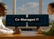 Two professionals sit at a wooden table facing dual monitors displaying IT diagrams with a bold 'Co-Managed IT' banner across the center (informative).