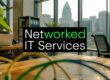 Office with a laptop on a wooden desk and a bold black sign that reads 'Networked IT Services' against a city skyline window backdrop