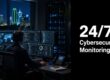 Security analyst at a dark, multi-monitor workstation monitoring cyber data, with a city skyline outside the window and a bold 24/7 Cybersecurity Monitoring sign to the right.