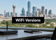 WiFi Versions banner over an office desk scene with a wireless router and notebook, overlooking a city skyline through a window.