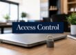 Laptop on a wooden desk with a small padlock in front and a dark blue 'Access Control' sign in the foreground, office setting behind.