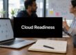 Office scene with a laptop, notebook, and mug on a wooden table; a black slide reading 'Cloud Readiness' is centered on screen, suggesting a presentation about cloud strategy.