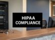 Reception desk with a black sign reading 'HIPAA COMPLIANCE' and a stethoscope on the counter, office servers in the background.
