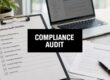 Compliance audit checklist on a clipboard with a laptop in the background.