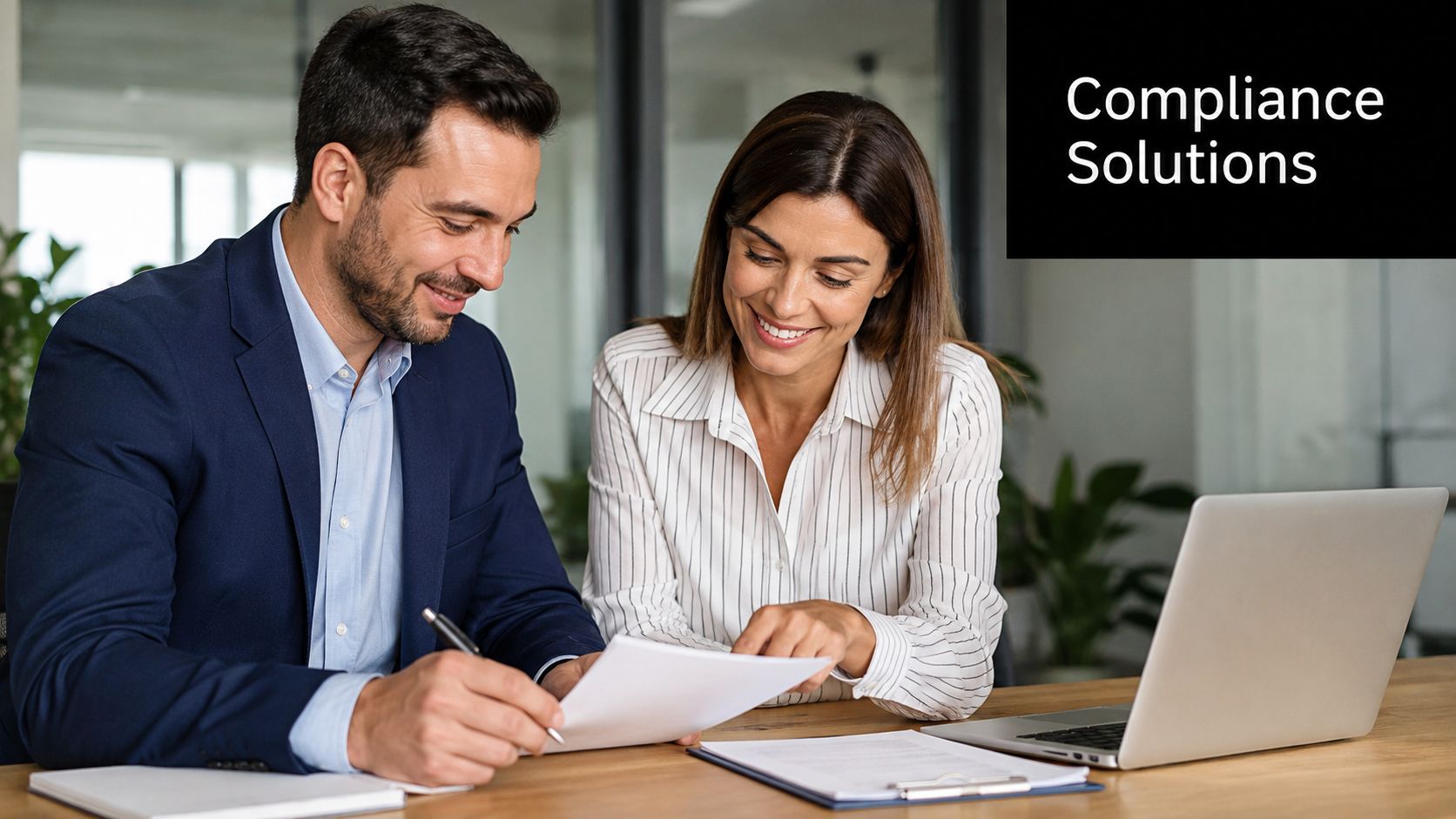 A professional man and woman smiling while reviewing documents together at an office desk with a laptop.