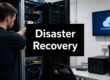 Data center racks with network cables and a large black sign reading 'Disaster Recovery' in the foreground, conveying IT recovery focus