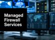 Black sign reading 'Managed Firewall Services' in a data-center control room with multiple monitors showing maps and charts nearby.
