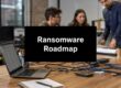 Sign reading 'Ransomware Roadmap' on a workstation with a laptop, cables, and a coffee cup; coworkers discuss in the background.