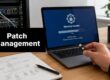 Laptop screen shows 'Security Update' progress while a hand holds a USB drive; 'Patch Management' sign on the desk.