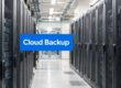 Data center corridor with rows of server racks and a blue sign reading 'Cloud Backup' across the aisle.